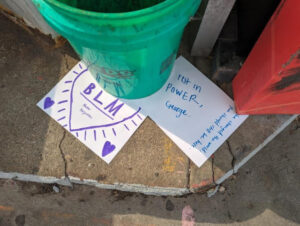 A green bucket with two pieces of paper that say "BLM" with a heart and "Rest in Power, George"